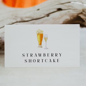 May include: A white card with a hand-drawn illustration of a beer glass and a champagne flute. The text "STRAWBERRY SHORTCAKE" is printed in black below the illustration.