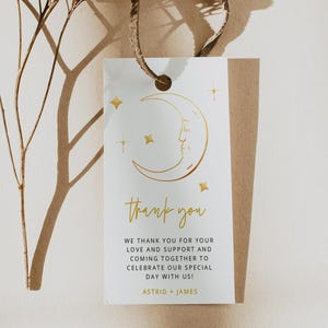 Puede incluir: Una etiqueta de agradecimiento blanca con un diseño de luna creciente y estrellas doradas. El texto dice "gracias" y "¡Les agradecemos su amor y apoyo y por reunirse para celebrar nuestro día especial con nosotros! Astrid + James".