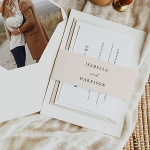 May include: A wedding invitation suite with a blush pink ribbon and a photo of a couple in a field. The invitation reads "By Isabella and Harrison".