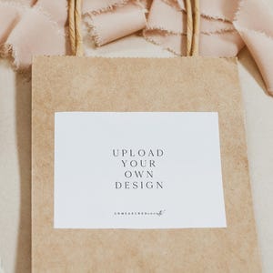 May include: A brown paper gift bag with a white square label that says "UPLOAD YOUR OWN DESIGN" and the website address "unmeasuredevent.com". The bag is sitting on a light brown surface with a pink ribbon.