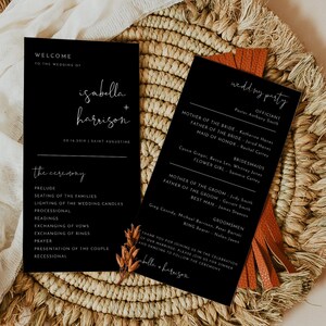 Puede incluir: Tarjetas negras de ceremonia y recepción de boda con texto en caligrafía blanca. Las tarjetas están sobre una estera de paja tejida con una borla marrón.