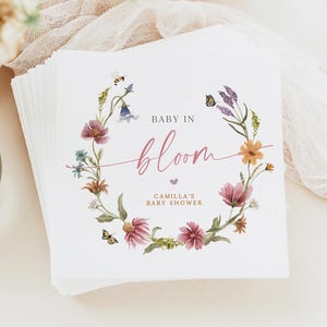 May include: A stack of square white baby shower invitations. Each invitation features a floral wreath design with the words "Baby in Bloom" in pink script. The text "Camilla's Baby Shower" is also included.