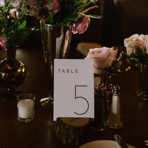 Puede incluir: Una tarjeta blanca con texto negro que dice "TABLE 5" se encuentra sobre una base de madera. La tarjeta está rodeada de flores rosas y blancas en jarrones de plata y vidrio.