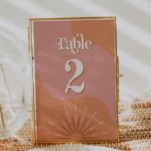 May include: A framed table number card with a gold border. The card features the word "Table" and the number "2" in a decorative font. The background has a retro design with shades of pink and orange, and a sunburst graphic.