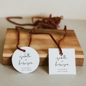 May include: Two white wedding favor tags with brown string. One tag is round and the other is rectangular. Both tags have the text "isabella + harrison" and the date "07.08.2019" with the location "NAPA, FLORIDA" printed below.