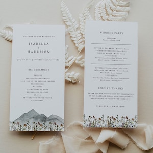 Könnte beinhalten: Zwei Hochzeitskarten mit Aquarell-Illustrationen. Die linke Karte zeigt eine Bergkette mit Wildblumen und den Text "Willkommen zur Hochzeit von Isabella + Harrison, 7. Juli 2021, Breckenridge, Colorado, Die Zeremonie, Präludium, Platzierung der Familien, Anzünden der Hochzeitskerzen, Einzug, Lesung, Austausch der Gelübde, Austausch der Ringe, Gebet, Vorstellung des Paares, Auszug". Die rechte Karte hat einen floralen Rand und den Text "Hochzeitsgesellschaft, Standesbeamter, Mutter der Braut, Vater der Braut, Brautjungfer, Brautjungfern, Blumenmädchen, Mutter des Bräutigams, Vater des Bräutigams, Trauzeuge, Trauzeugen, Ringträger, Besonderer Dank, Vielen Dank, dass Sie an unserer Hochzeitsfeier teilgenommen haben. Bitte schließen Sie sich uns zum Abendessen und Tanzen an, das nach der Zeremonie stattfindet, Isabella + Harrison".