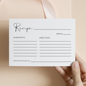 May include: A white recipe card with the word "Recipe" in elegant script. The card includes spaces for ingredients, directions, and servings. A light pink envelope with a matching ribbon is in the background. The card is held by a hand.