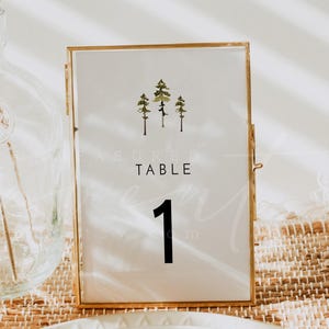 May include: A gold-framed table number card with a white background. The card features a watercolor illustration of three green trees and the text "TABLE 1".