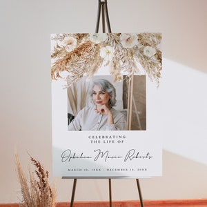 Puede incluir: Un cartel conmemorativo blanco con una foto de una mujer y el texto "Celebrating the life of Ophelia Maria Roberts March 05, 19XX - December 18, 20XX". El cartel está decorado con flores secas y hierba de la pampa.
