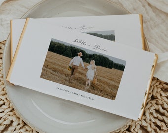 May include: A white wedding invitation with a gold border. The invitation features a photo of a couple walking in a field, with the text "Ella + Harrison" and "08.09.20XX | Saint Augustine" printed below the photo.