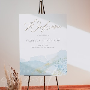 Może przedstawiać: Tablica powitalna na ślub z niebieskim i złotym tłem akwarelowym. Na tablicy widnieje napis "Witamy na ślubie Izabeli + Harrisona, 7 lipca 2019, Saint Augustine, Floryda".