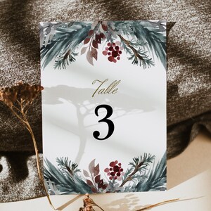 May include: A white card with a watercolor design of pine branches and red berries. The card says "Table 3" in gold lettering.