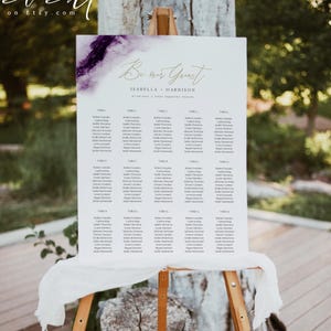 May include: A wedding seating chart on a wooden easel. The white sign features a purple and gold watercolor design at the top, with the words "Be our Guest" in elegant script. The names of the wedding couple and guests are listed below.