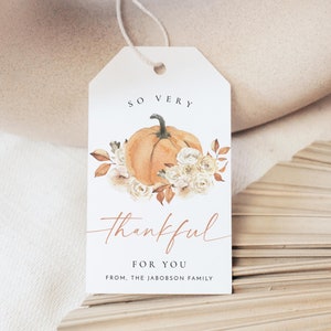 Puede incluir: Una etiqueta de regalo blanca con una ilustración de acuarela de una calabaza y flores blancas. El texto en la etiqueta dice "So Very Thankful For You From, The Jacobson Family."