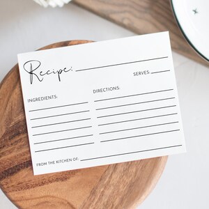 May include: A white recipe card with the word "Recipe" in cursive script, placed on a wooden bowl. A white plate with black cross patterns and a rolled-up striped towel are in the background.