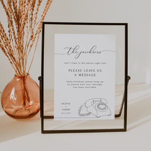 May include: A white sign with black text in a black frame. The sign says "the jacobsons can't come to the phone right now. Please leave us a message. When finished, please hang up to ensure that your message has been recorded." Below the text is a line drawing of a vintage rotary phone. The sign also includes the names "Olivia & Asher" and the date "08.18.2013".