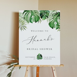 May include: A white sign with green tropical leaves at the top. The sign says "Welcome to Hannah's Bridal Shower" with the date "September 08" and location "Palm Beach, Florida".