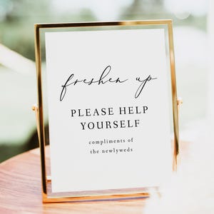 May include: A gold-framed sign with the text "freshen up" in a cursive font. Below the text is "PLEASE HELP YOURSELF" in a sans-serif font. The bottom of the sign reads "compliments of the newlyweds".