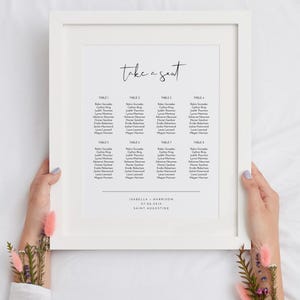Puede incluir: Un diagrama de asientos de boda blanco enmarcado con el texto "take a seat" en la parte superior. El diagrama enumera las mesas con los nombres de los invitados para una recepción de boda. La parte inferior del diagrama incluye los nombres "Isabella & Harrison", la fecha "07.08.2019" y la ubicación "Saint Augustine".