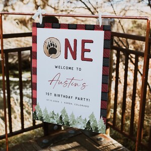 May include: A white sign with red and black buffalo plaid border. The sign says "ONE" in red letters with a brown bear paw print in a wood circle. The text below says "Welcome to Austin's 1st Birthday Party! 07.15.20XX | ASPEN, COLORADO".