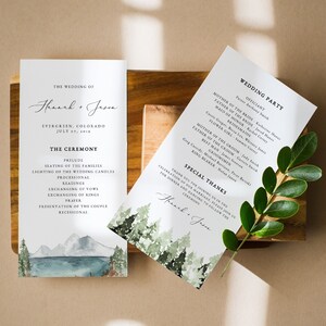 May include: Wedding ceremony program with a watercolor illustration of a mountain range and a lake. The program lists the order of events for the ceremony, including the seating of the families, lighting of the candles, and the exchanging of vows. The program also includes the names of the wedding party.