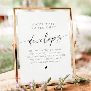 Puede incluir: Un cartel blanco con texto negro que dice "Can't wait to see what develops."  El cartel también incluye instrucciones para usar una cámara para capturar momentos especiales en un evento.  El cartel está en un marco dorado y está sentado en una mesa con flores.