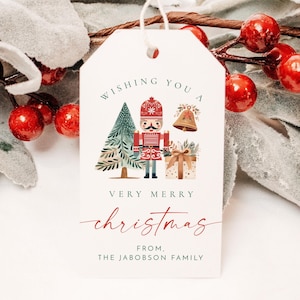 May include: A white gift tag with a red and green illustration of a nutcracker holding a present and standing in front of a Christmas tree. The text on the tag reads "Wishing you a very merry christmas from the Jacobson family."