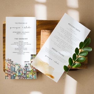 Puede incluir: Programa de la ceremonia de boda con una ilustración de acuarela de una calle de la ciudad. El programa enumera el orden de los eventos de la ceremonia, incluyendo la colocación de las familias, el encendido de las velas de la boda y la procesión. El programa también incluye los nombres de la fiesta de bodas y una sección de agradecimiento especial.