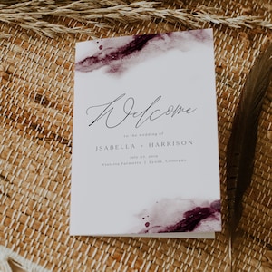 May include: A white wedding welcome sign with a watercolor background in shades of purple. The text reads "Welcome to the wedding of Isabella + Harrison July 07, 2019 Violetta Farmette | Lyons, Colorado".