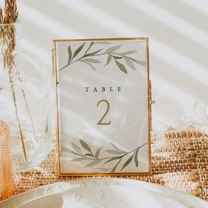 Peut inclure: Carte de numéro de table avec un cadre doré et un motif aquarelle de feuilles vertes et le texte "TABLE 2" sur fond blanc.