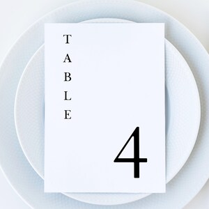 Peut inclure: Une carte de table blanche avec le mot "TABLE" imprim&eacute; verticalement et le chiffre "4" en noir. La carte est plac&eacute;e sur une assiette blanche avec un bord textur&eacute;, adapt&eacute;e &agrave; un mariage ou &agrave; un &eacute;v&eacute;nement formel.