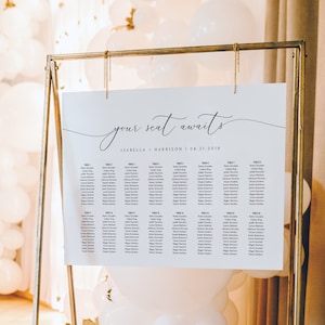 Puede incluir: Un plano de asientos de boda blanco con el texto "your seat awaits" en escritura cursiva negra. El gráfico enumera los nombres de los invitados por número de mesa. El gráfico se muestra en un caballete de metal dorado con globos blancos detrás.