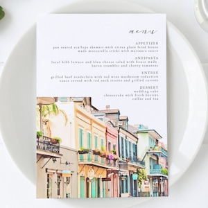 May include: A watercolor illustration of a New Orleans street scene with colorful buildings, balconies, and flowers. The menu features a selection of appetizers, antipasti, entrees, and desserts. The text reads: "menu", "appetizer", "pan seared scallops skewers with citrus glaze fried house made mozzarella sticks with marinara sauce", "antipasta", "local bibb lettuce and bleu cheese salad with house made bacon crumbles and cherry tomatoes", "entree", "grilled beef tenderloin with red wine mushroom reduction sauce served with red neck risotto and grilled carrots", "dessert", "wedding cake", "cheesecake with fresh berries", "coffee and tea".