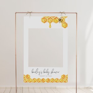 May include: A white photo booth backdrop featuring a honeycomb and bee design. The backdrop has a cutout for a person's face. The text "bailey's baby shower" and "AUGUST 06, 2020" is printed on the backdrop.