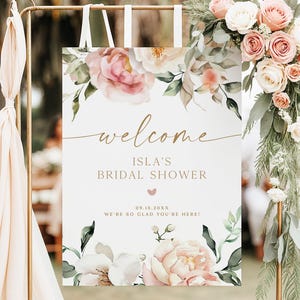 以下が含まれることがあります： イラさんのブライダルシャワー用のウェルカムサイン。ピンクと白のバラと緑の葉のフローラルデザイン。サインは金色の金属スタンドにあり、日付と歓迎のメッセージが含まれています。