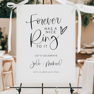 May include: A white sign on a black easel with the text "Forever has a nice ring to it." Below, it reads "Let's celebrate Jolie + Michael, we're so glad you're here." White ribbons are tied to the top.