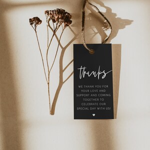 May include: A black gift tag with the word "thanks" in elegant white script. The tag includes a message of gratitude and is tied with a natural twine. Dried flowers and their shadows add a delicate touch.