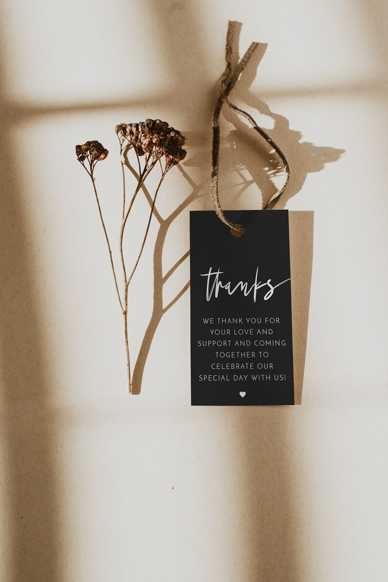May include: A black gift tag with the word "thanks" in elegant white script. The tag includes a message of gratitude and is tied with a natural twine. Dried flowers and their shadows add a delicate touch.