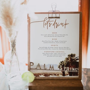 May include: A white sign with brown text that reads "let's drink" and lists various beer, wine, and spirit options. The sign is propped up on a wooden stand with a picture of a desert landscape and the words "Palm Springs" in large letters.