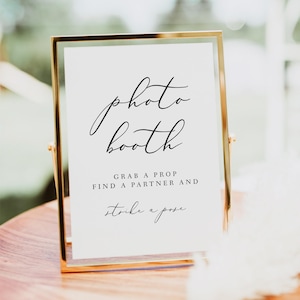 May include: A gold-framed sign with white background and black text that reads "photo booth" in cursive. Below the text, it says "Grab a prop, find a partner and strike a pose."