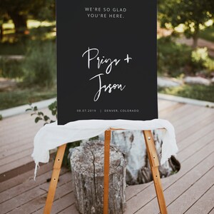 May include: A black wedding sign on a wooden easel. The sign reads "WE'RE SO GLAD YOU'RE HERE." and features the names "Prigr + Jison" and the date and location "08.07.2019 | DENVER, COLORADO" in white script.