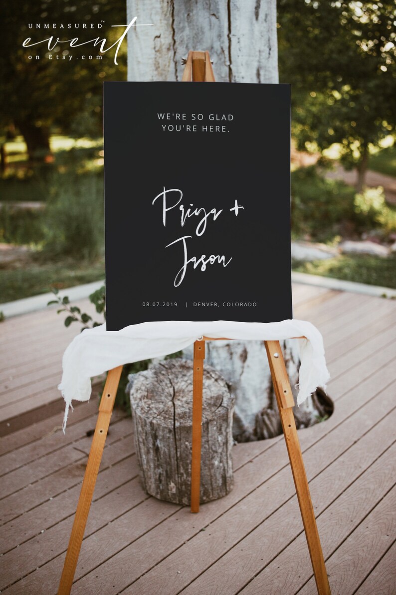 May include: A black wedding sign on a wooden easel. The sign reads "WE'RE SO GLAD YOU'RE HERE." and features the names "Prigr + Jison" and the date and location "08.07.2019 | DENVER, COLORADO" in white script.