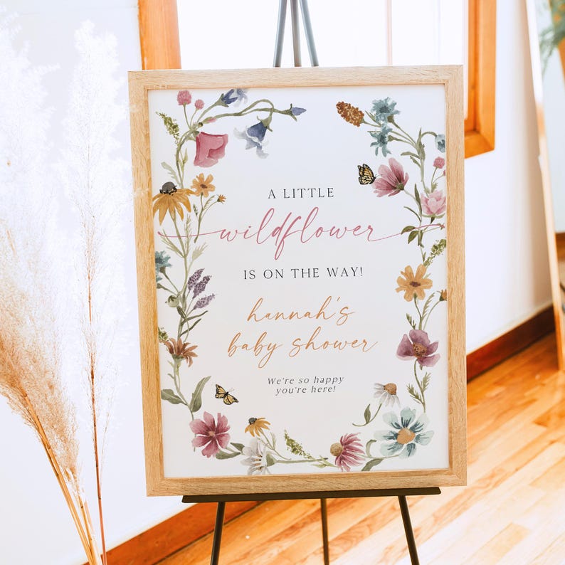 May include: A white sign with a floral border and the text "A little wildflower is on the way! Hannah's baby shower. We're so happy you're here!"