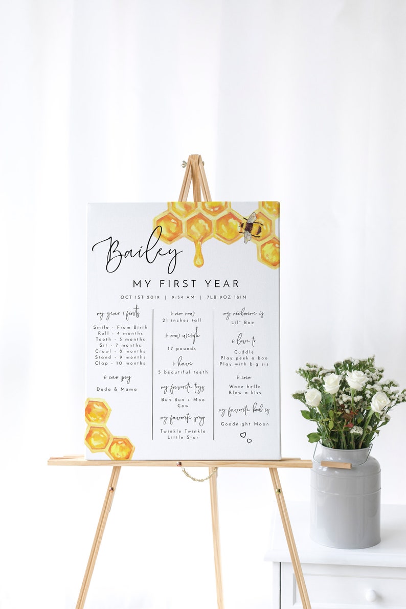 May include: A white poster board with a honey comb design and a bee. The poster board has the text "Bailey My First Year" and lists milestones for a baby's first year, including weight, height, first words, and favorite things.