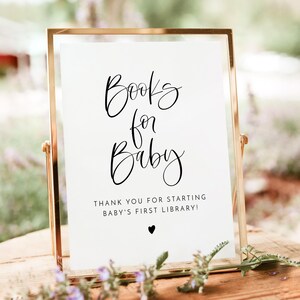 May include: A white sign with black calligraphy that reads "Books for Baby" and "Thank you for starting baby's first library!" with a small black heart at the bottom. The sign is in a gold frame.
