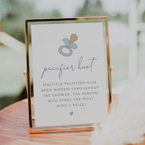 May include: A white sign with a gold frame, announcing a pacifier hunt. The sign has a blue and brown illustration of a pacifier and the text "pacifier hunt". The text below the illustration reads "Multiple pacifiers have been hidden throughout the shower. The person who finds the most wins a prize!"
