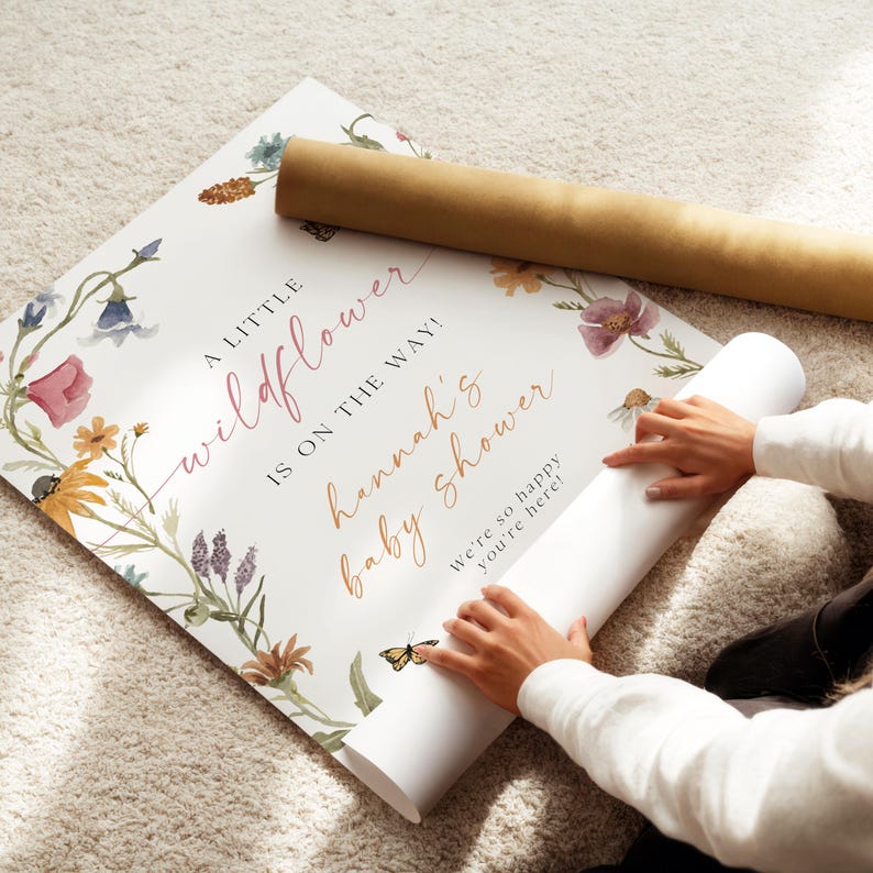 May include: A white poster with a floral design and the text "A little wildflower is on the way! Hannah's baby shower. We're so happy you're here!"