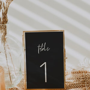 May include: A black table number card in a gold frame, displaying the word "table" and the number "1" in white script. The card is set on a woven placemat, with a vase of dried flowers in the background.