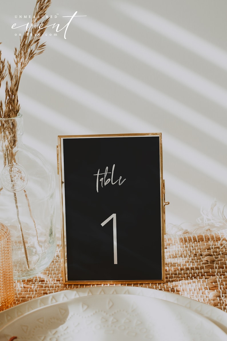 May include: A black table number card in a gold frame, displaying the word "table" and the number "1" in white script. The card is set on a woven placemat, with a vase of dried flowers in the background.