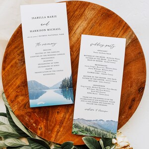 May include: Wedding ceremony programs with watercolor landscape art. The programs include the names of the couple, the wedding party, and the ceremony details. The programs are on a wooden surface with greenery.